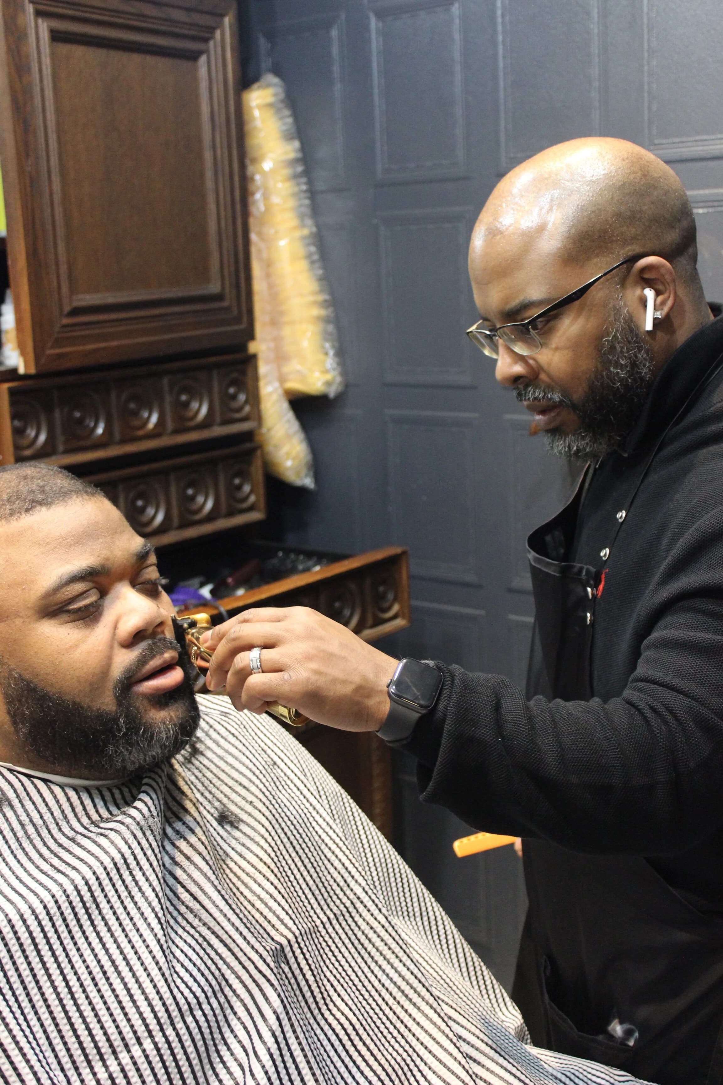 Barber with glasses uses gold trimmers to groom a man's beard in a barbershop.