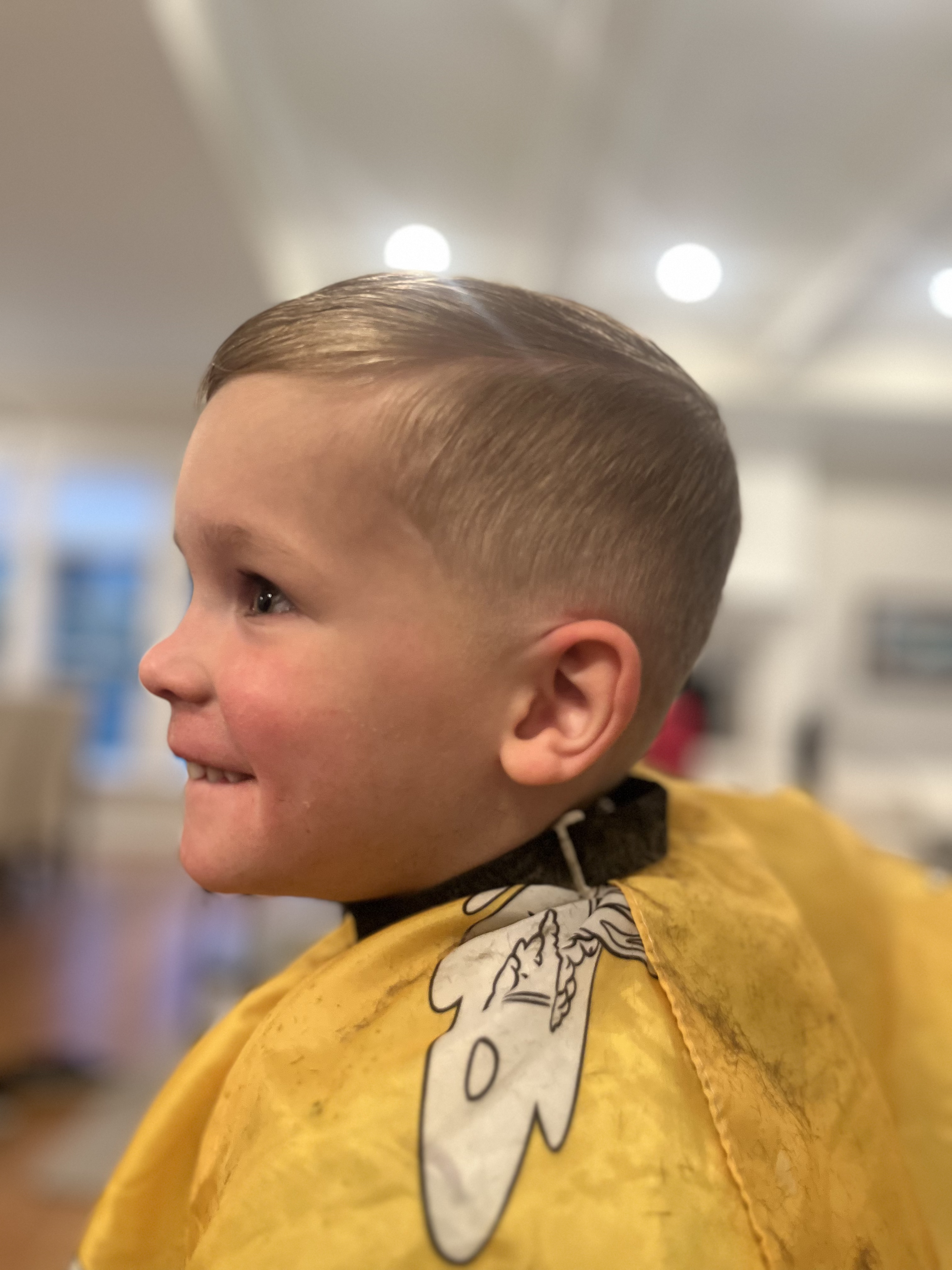 A smiling young boy with a fresh haircut wearing a yellow barber cape indoors.